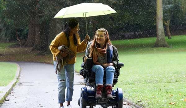 friends in the rain, one walking and one in an electric wheelchair, sharing an umbrella