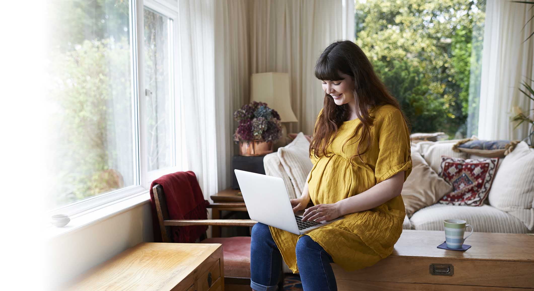 Pregnant person on sofa with cup of tea using laptop